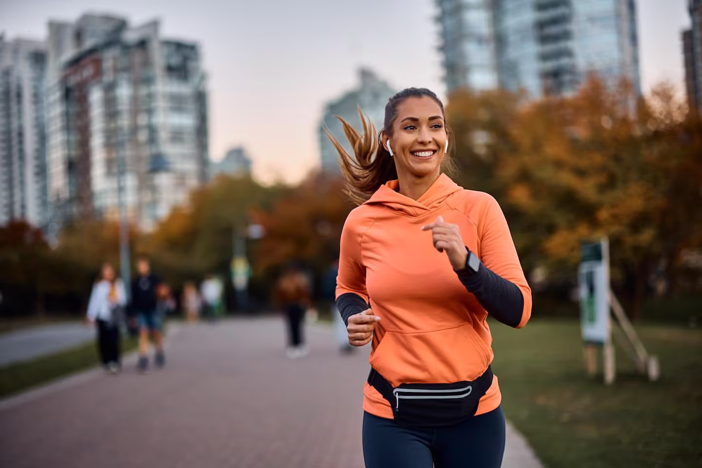 Smiling woman running outdoors in a city park wearing an orange hoodie, black leggings, and a black waist pouch.