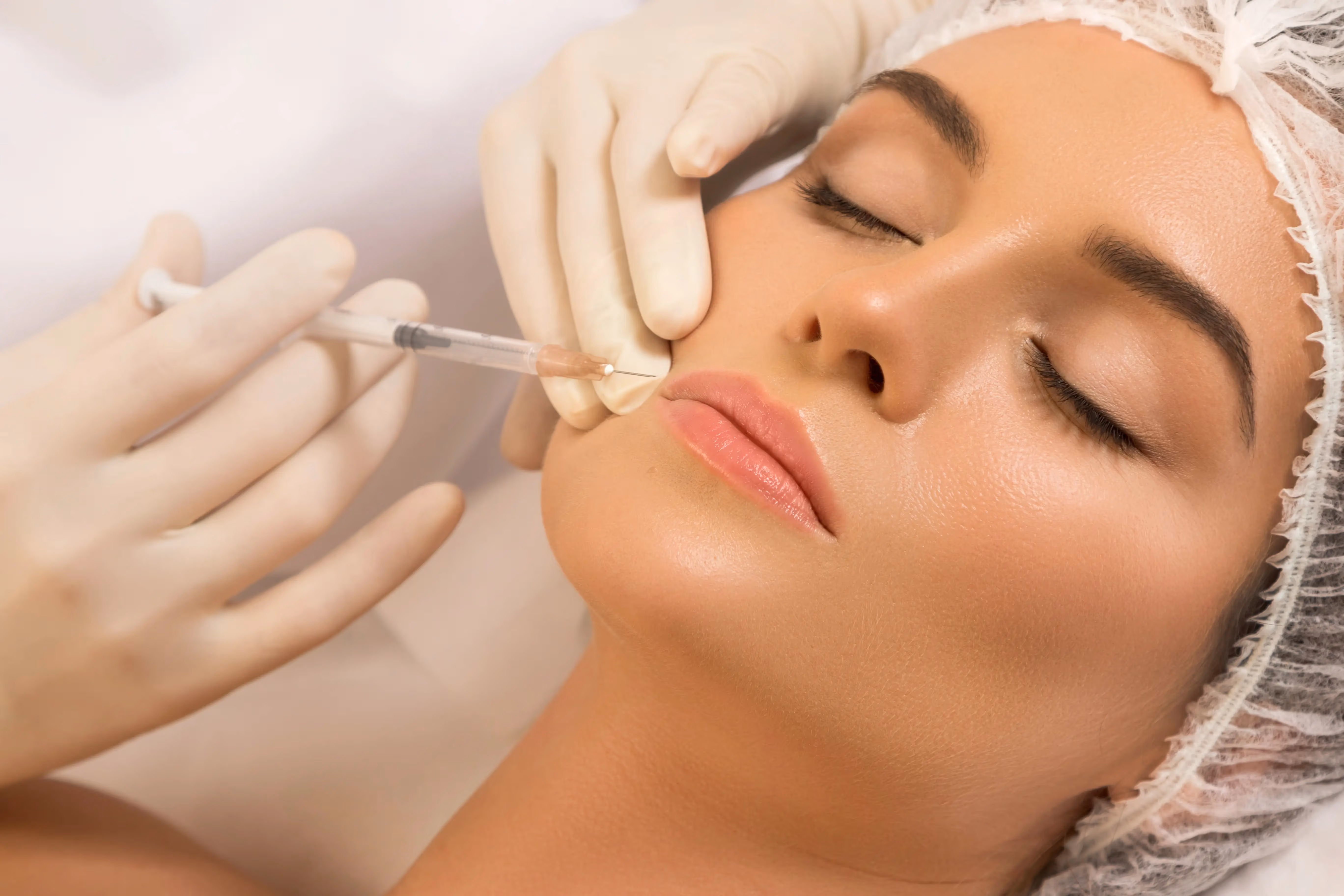 Close-up of a woman receiving a cosmetic injection in her lip while wearing a hair cover and with eyes closed.