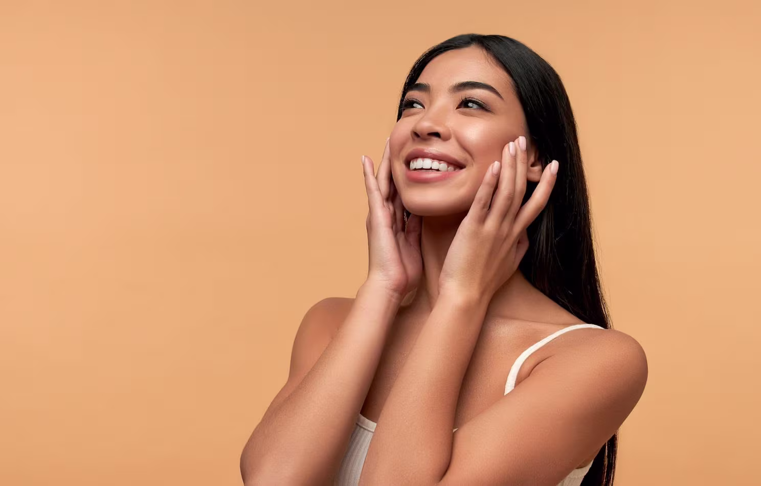 Smiling woman with long black hair touching her face against a light brown background.