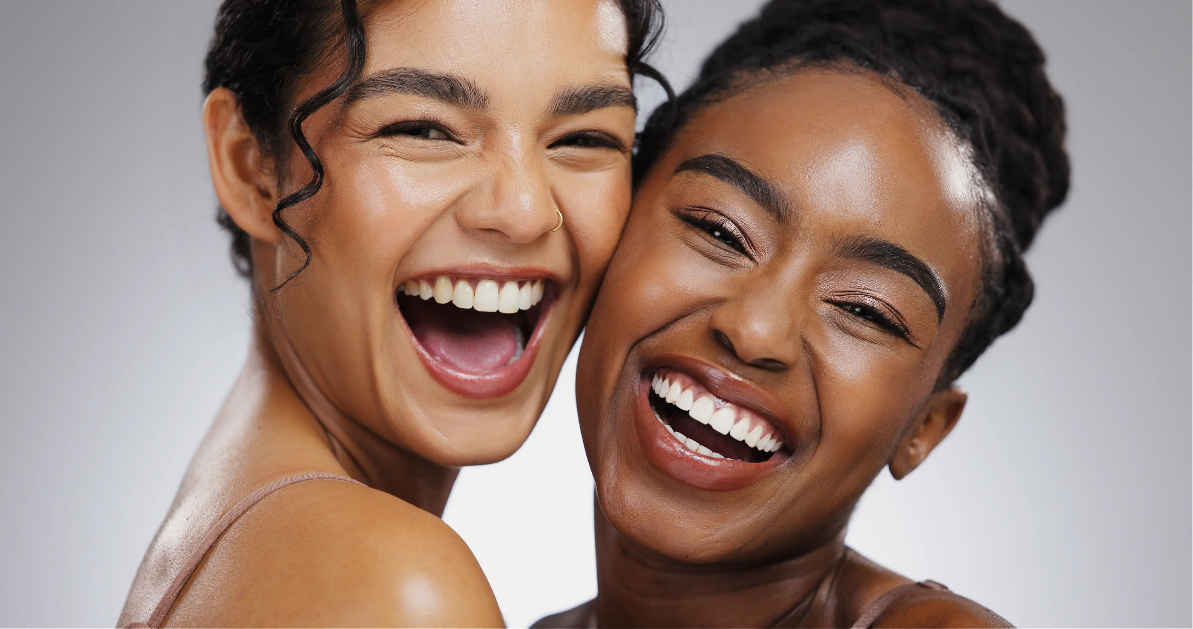 Two women smiling and laughing closely together against a neutral background.