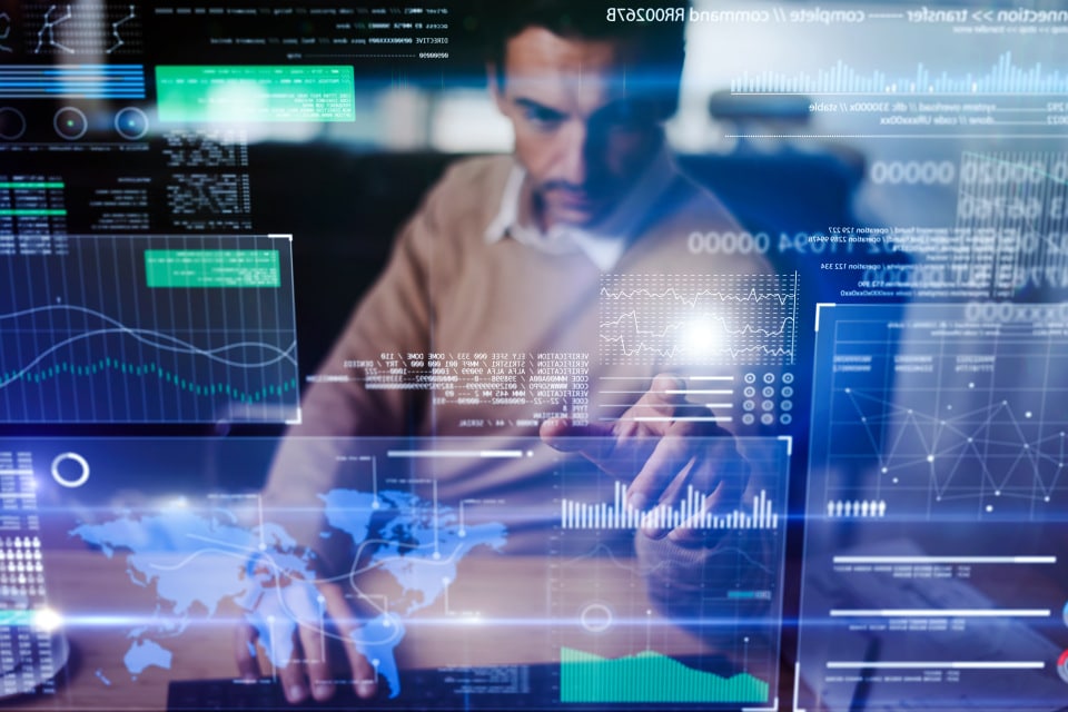 Man in a brown sweater working at a desk with a laptop, interacting with multiple transparent digital data screens showing graphs, charts, world maps, and data analytics overlays.