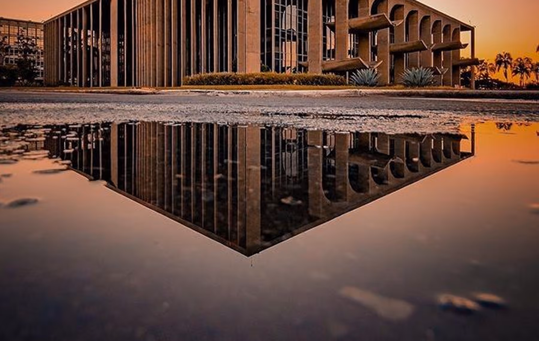 Palácio da Justiça em Brasília refletido em poça d'água ao pôr do sol com céu alaranjado e palmeiras ao fundo.