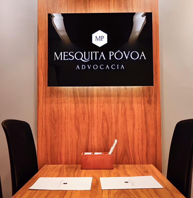 Sala de reunião com mesa de madeira, duas cadeiras pretas e logo da Mesquita Póvoa Advocacia na parede.