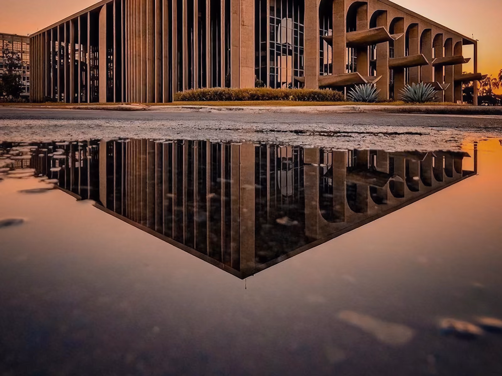 Palácio da Justiça em Brasília refletido em poça d'água ao pôr do sol com céu alaranjado e palmeiras ao fundo.
