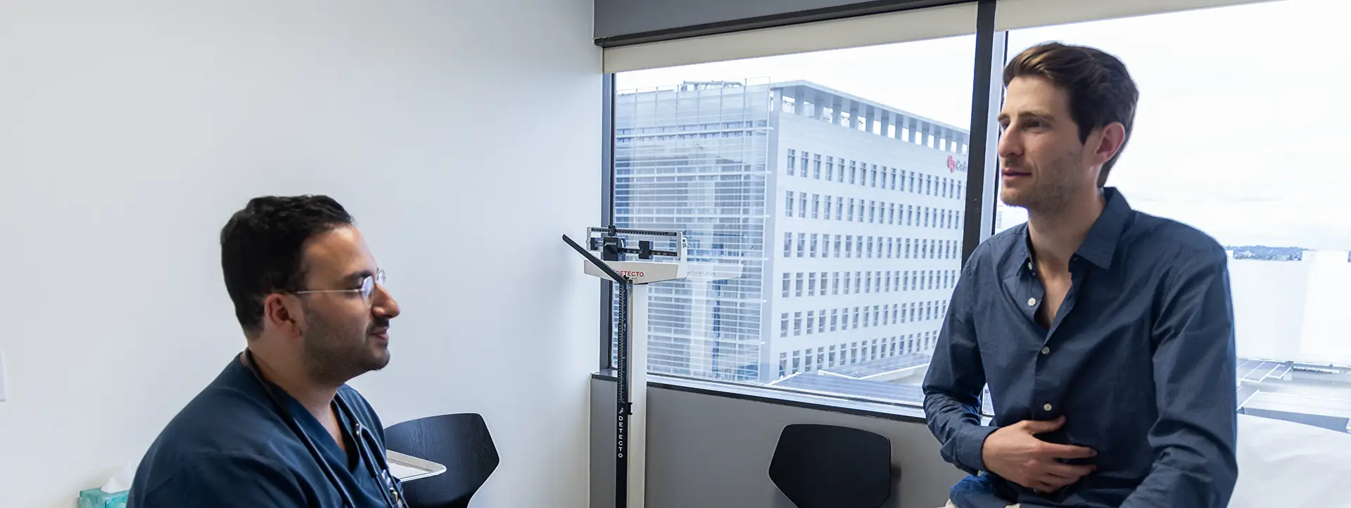 Doctor talking to a man holding his stomach in a medical office with a window view of a building.