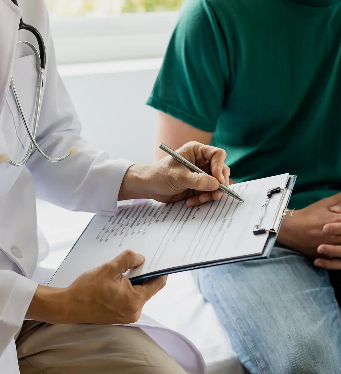Doctor holding clipboard and pen, writing notes while patient in green shirt sits nearby.
