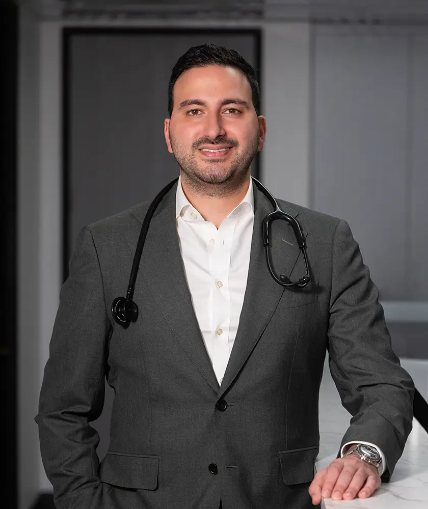 Smiling man in a grey suit with a stethoscope around his neck standing and leaning on a counter.