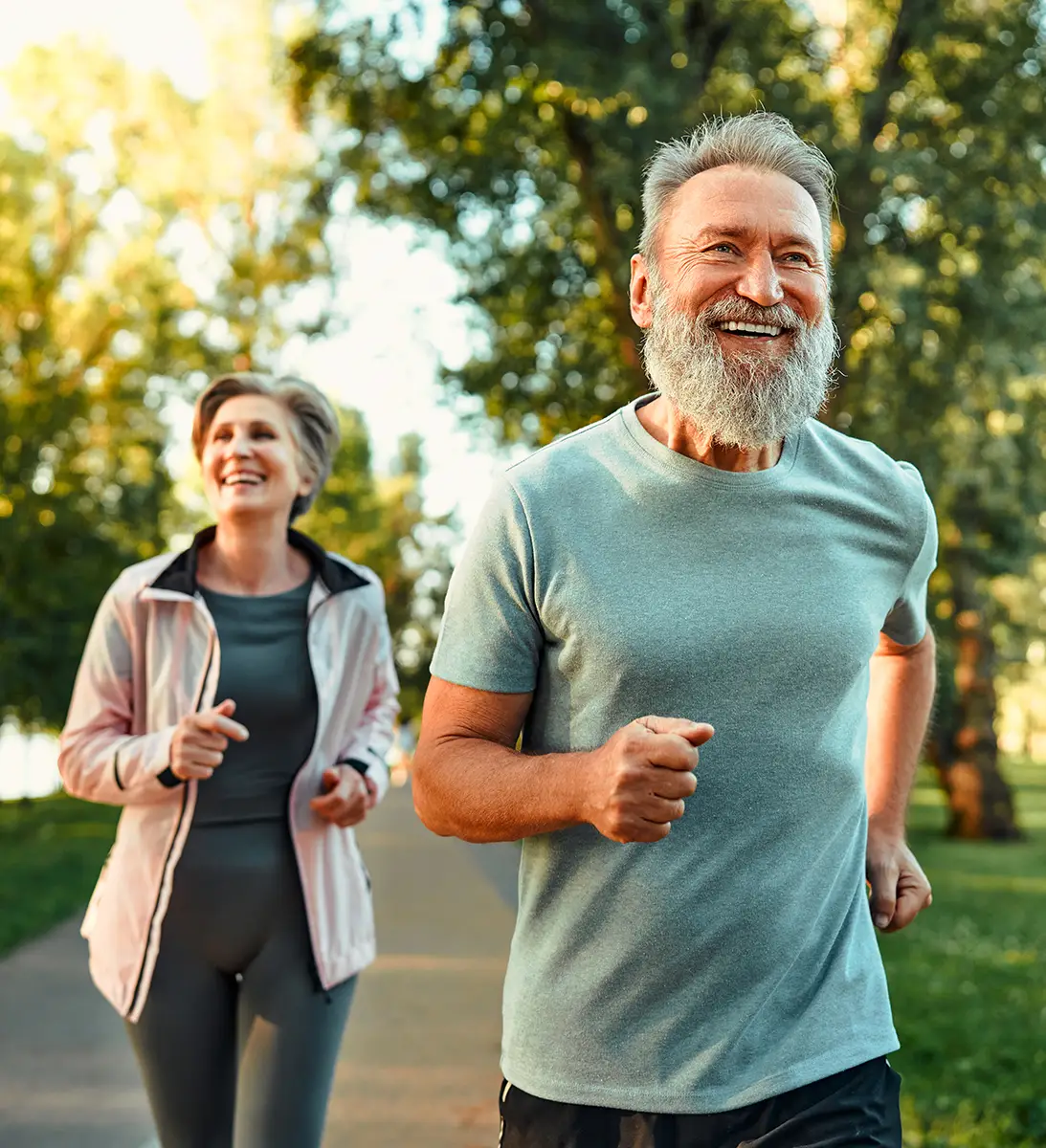 Smiling older man and woman jogging outside on a sunny day with trees in the background.