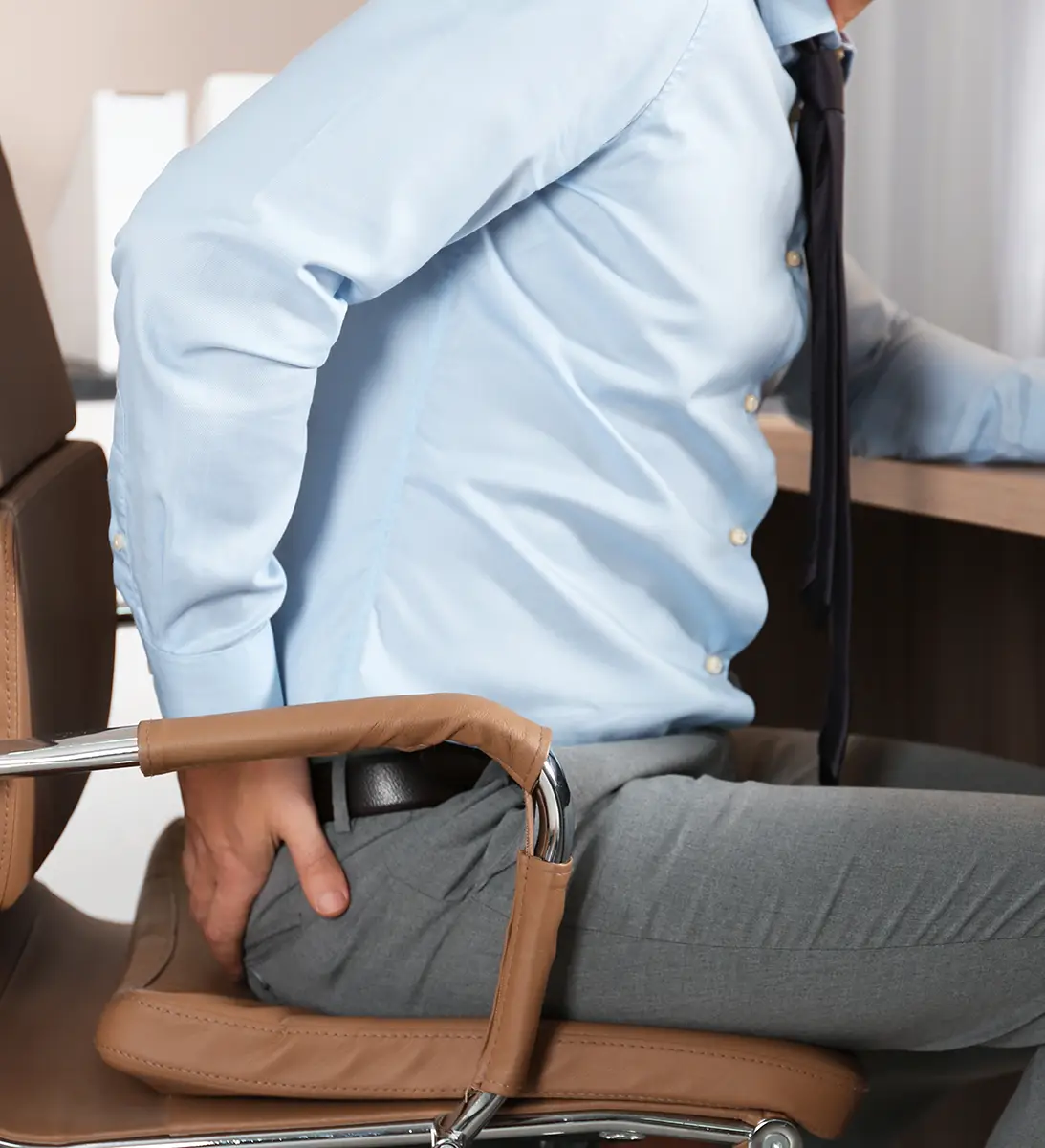 Man in office attire sitting on chair holding his lower back as if in pain or discomfort.