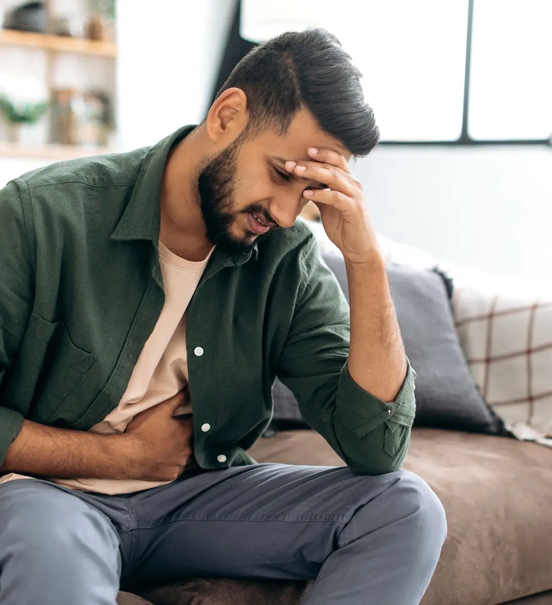 Man in green shirt sitting on couch holding stomach and forehead, showing discomfort or pain.
