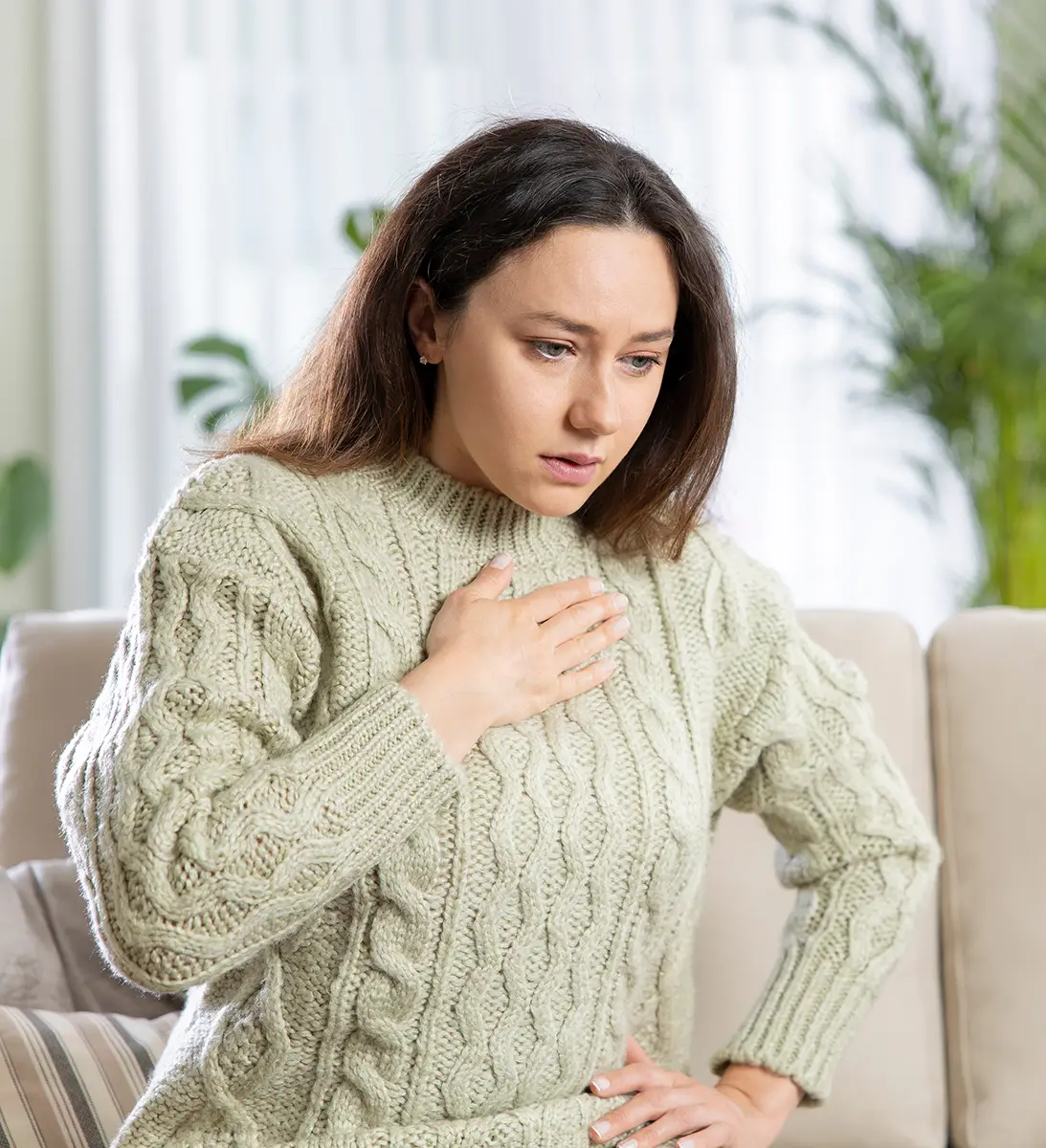 Woman sitting on a sofa clutching her chest with a concerned expression, wearing a knit sweater.
