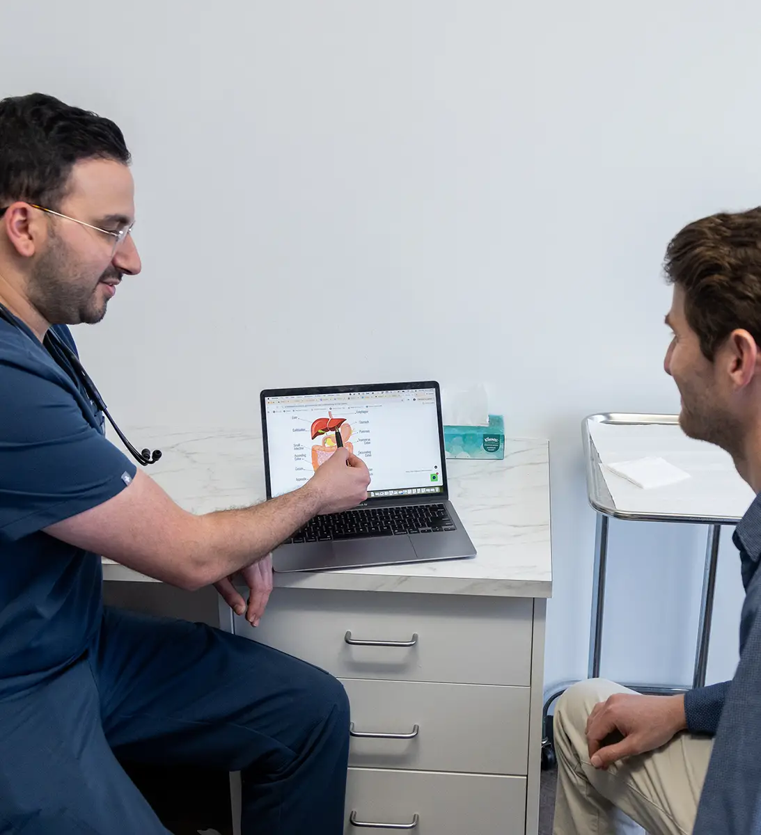 Doctor in scrubs showing a medical diagram on a laptop to a seated patient in a clinic room.