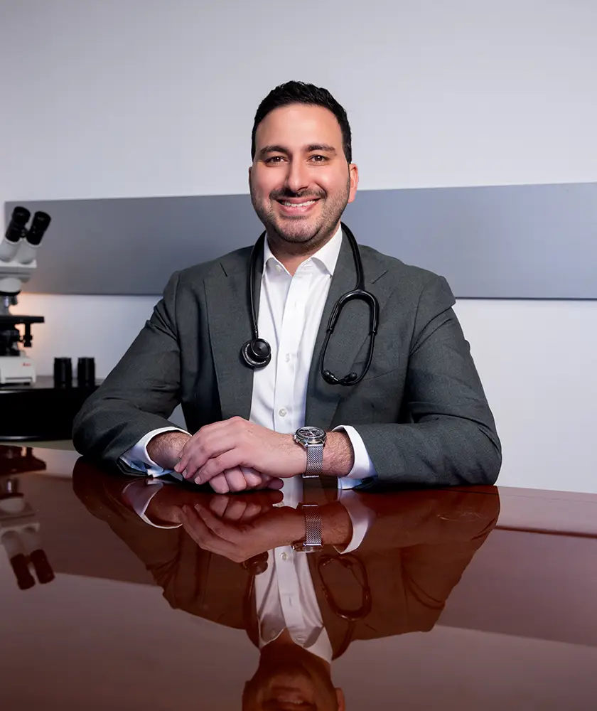 Smiling male doctor in a suit with a stethoscope around his neck sitting at a reflective desk.