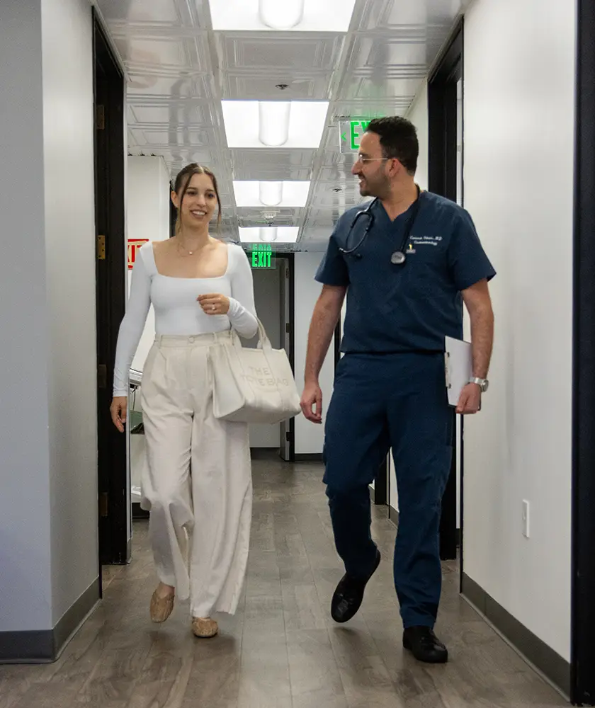 Woman in white top and pants walking and talking with a male doctor in blue scrubs in a hallway.