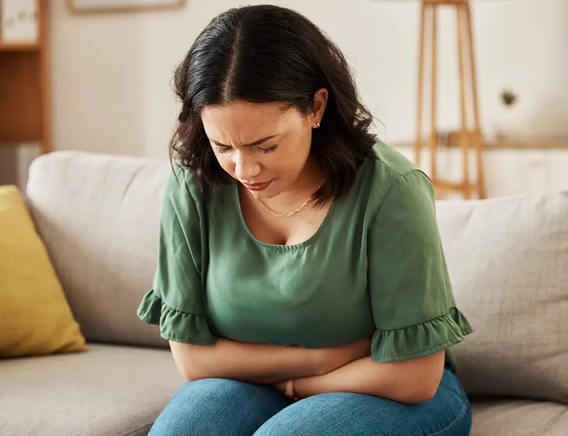 Woman sitting on couch with hands on stomach, appearing to have abdominal pain.