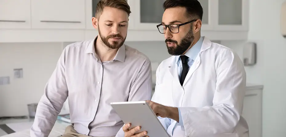 A doctor in a white coat shows information on a tablet to a man in a gray shirt in a medical office.