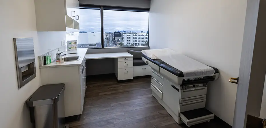 Modern medical exam room with an examination table, countertop with sink, and city view window.