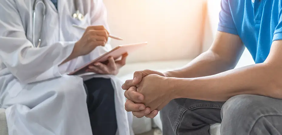 Doctor in white coat holding tablet, consulting patient with hands clasped in blue shirt and jeans.