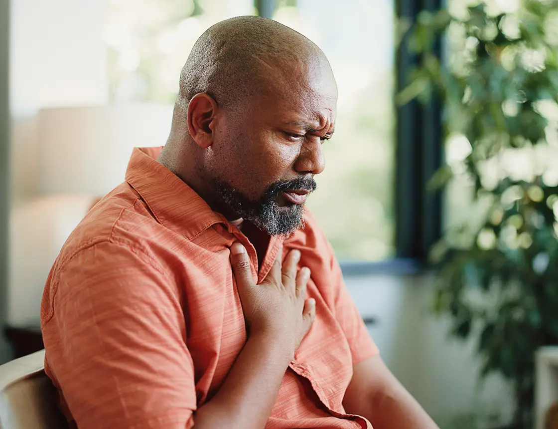 Man in orange shirt clutching his chest with a pained expression, sitting indoors near a window.