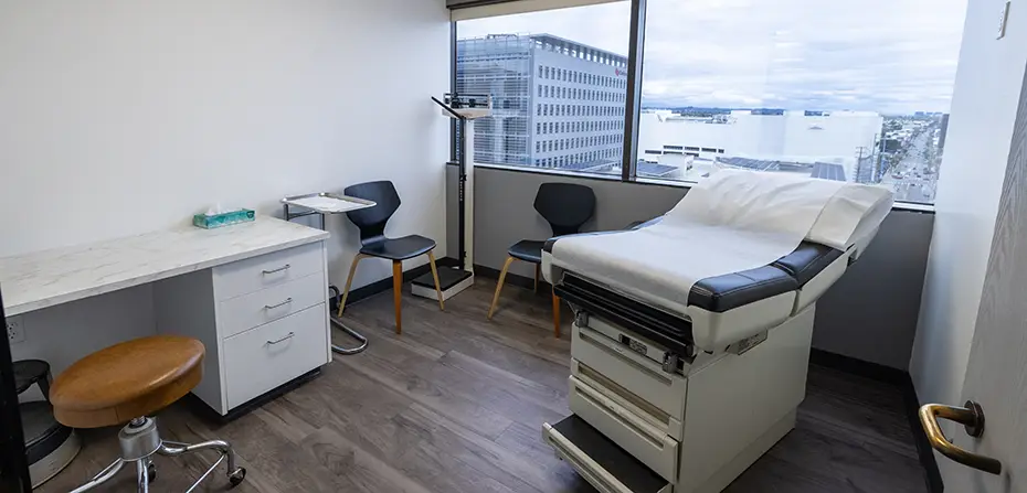 Medical exam room with patient table, chairs, scale, and large window overlooking city buildings.