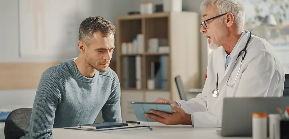 Male doctor in white coat shows tablet to a male patient seated at a desk in a consultation room.