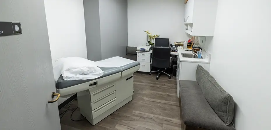 Small medical exam room with exam table, chair, desk, computer, and bench seating.