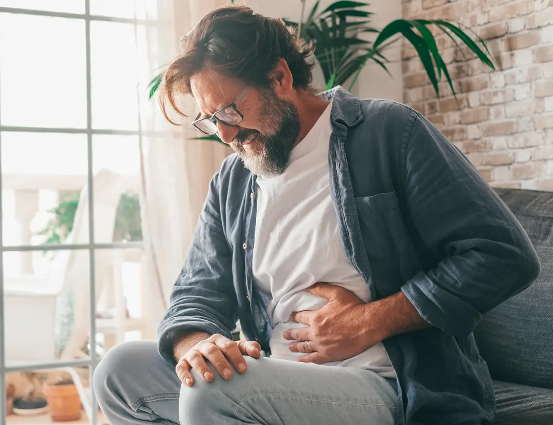 Middle-aged man sitting on a couch clutching his stomach in pain with a grimace on his face.