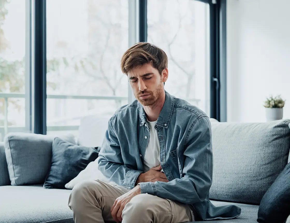 Man sitting on a sofa holding his stomach with a pained expression in a bright living room.