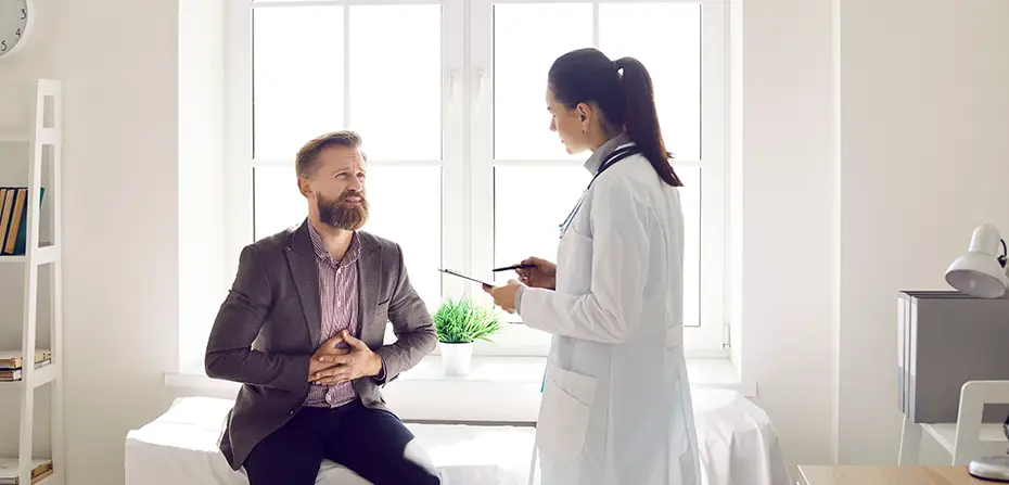 Man sitting on exam table clutching stomach, talking to female doctor with stethoscope in bright room.