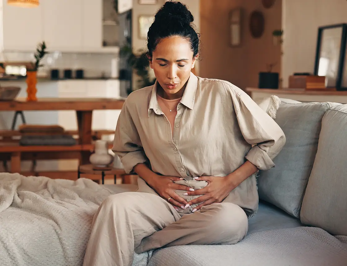 Woman sitting on sofa clutching her stomach, showing discomfort or pain.