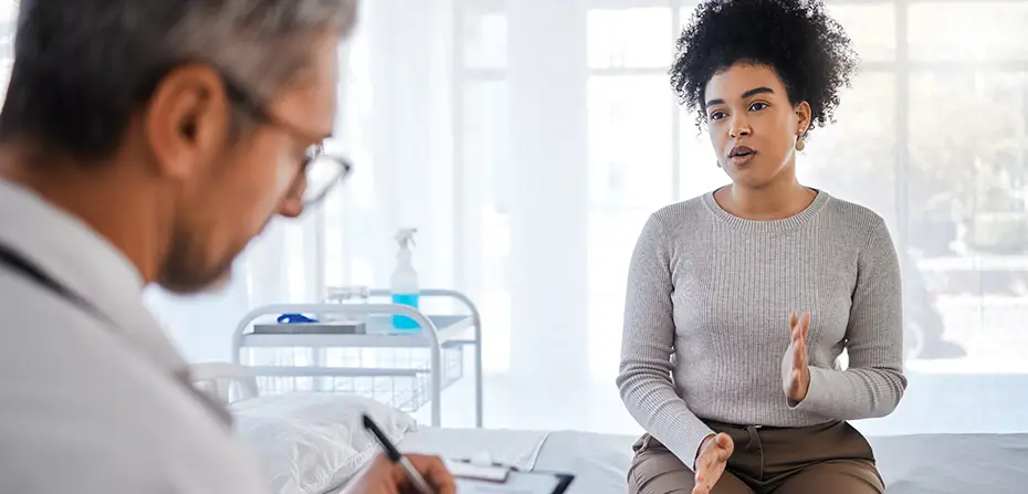 Woman explaining symptoms to a male doctor taking notes in a bright medical office.