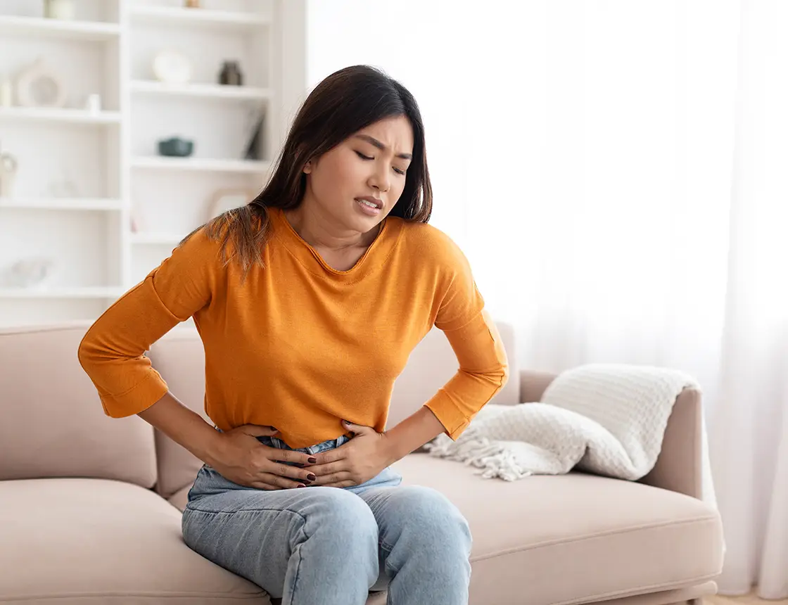 Woman in orange shirt sitting on a couch, clutching her abdomen in pain.