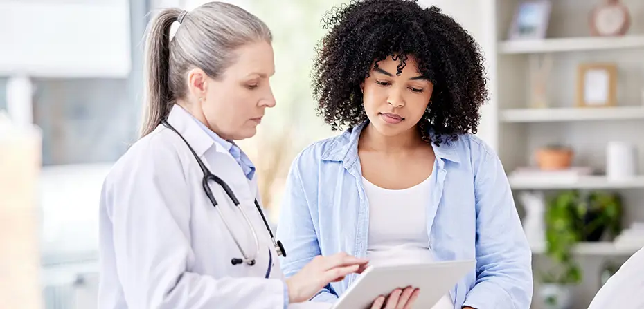 Doctor in white coat discussing medical information on tablet with woman in casual clothes.