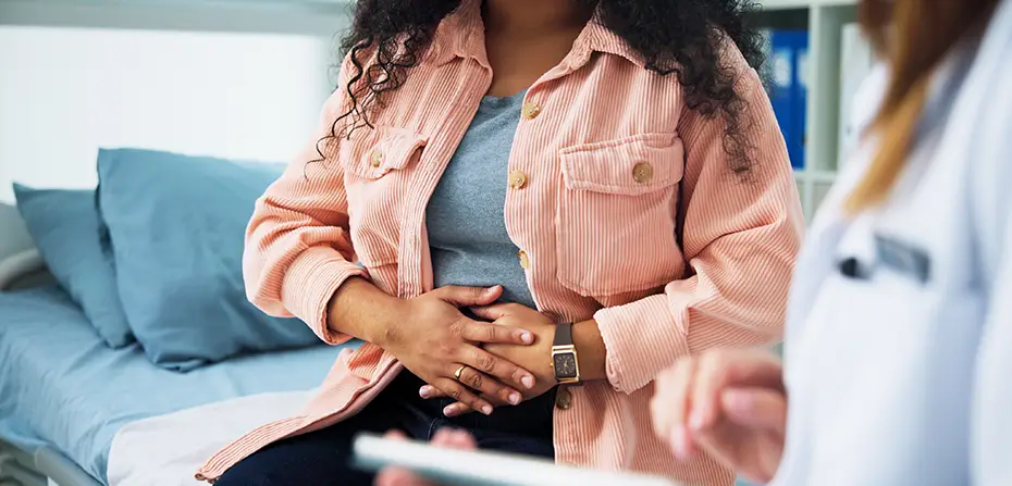 Woman in a peach jacket holding her stomach, sitting on a medical couch with a doctor nearby.