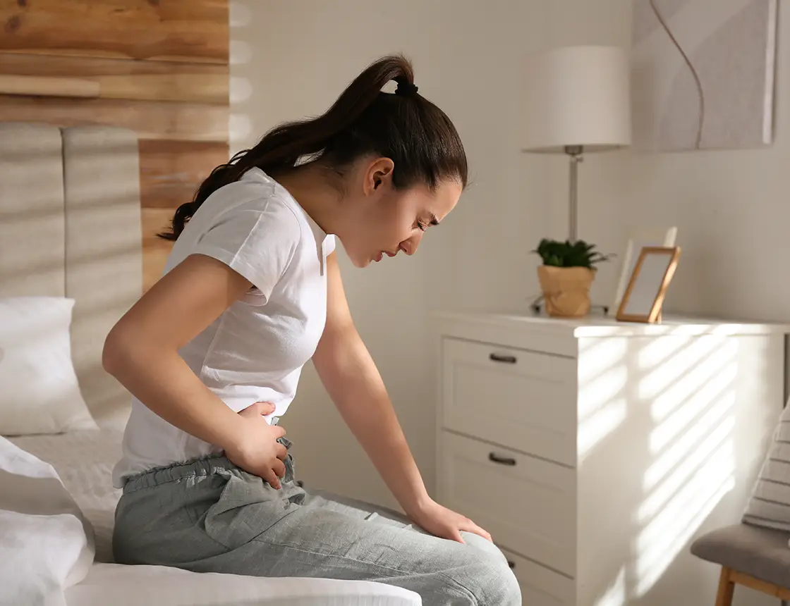 Woman sitting on bed, holding her stomach and wincing in pain in a softly lit bedroom.