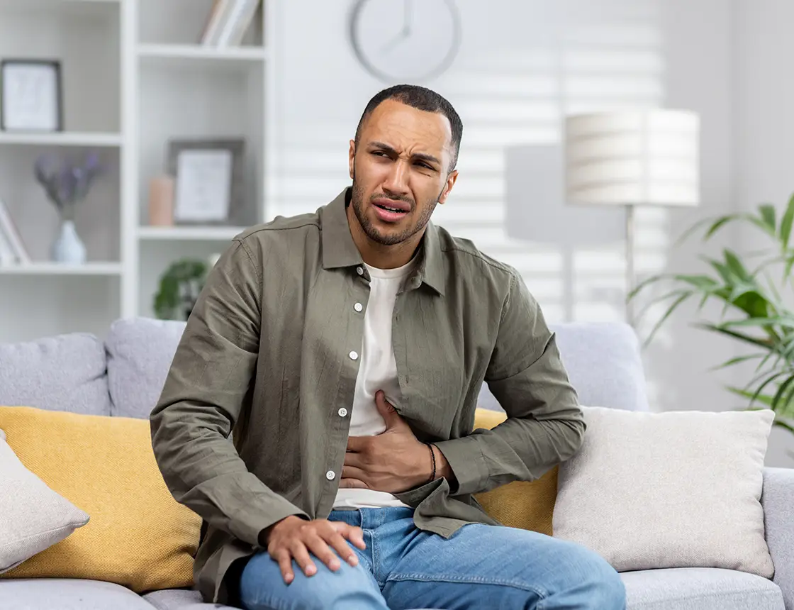 Man sitting on a couch holding his stomach with a pained expression in a bright living room.