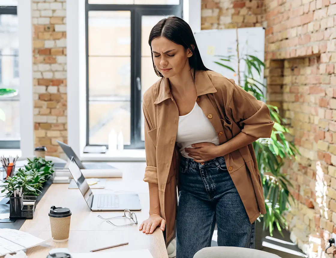 Woman in casual clothes standing by desk, clutching stomach with a pained expression in an office.