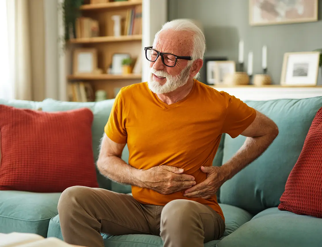 Elderly man in orange shirt sitting on a couch, holding his side with a pained expression.