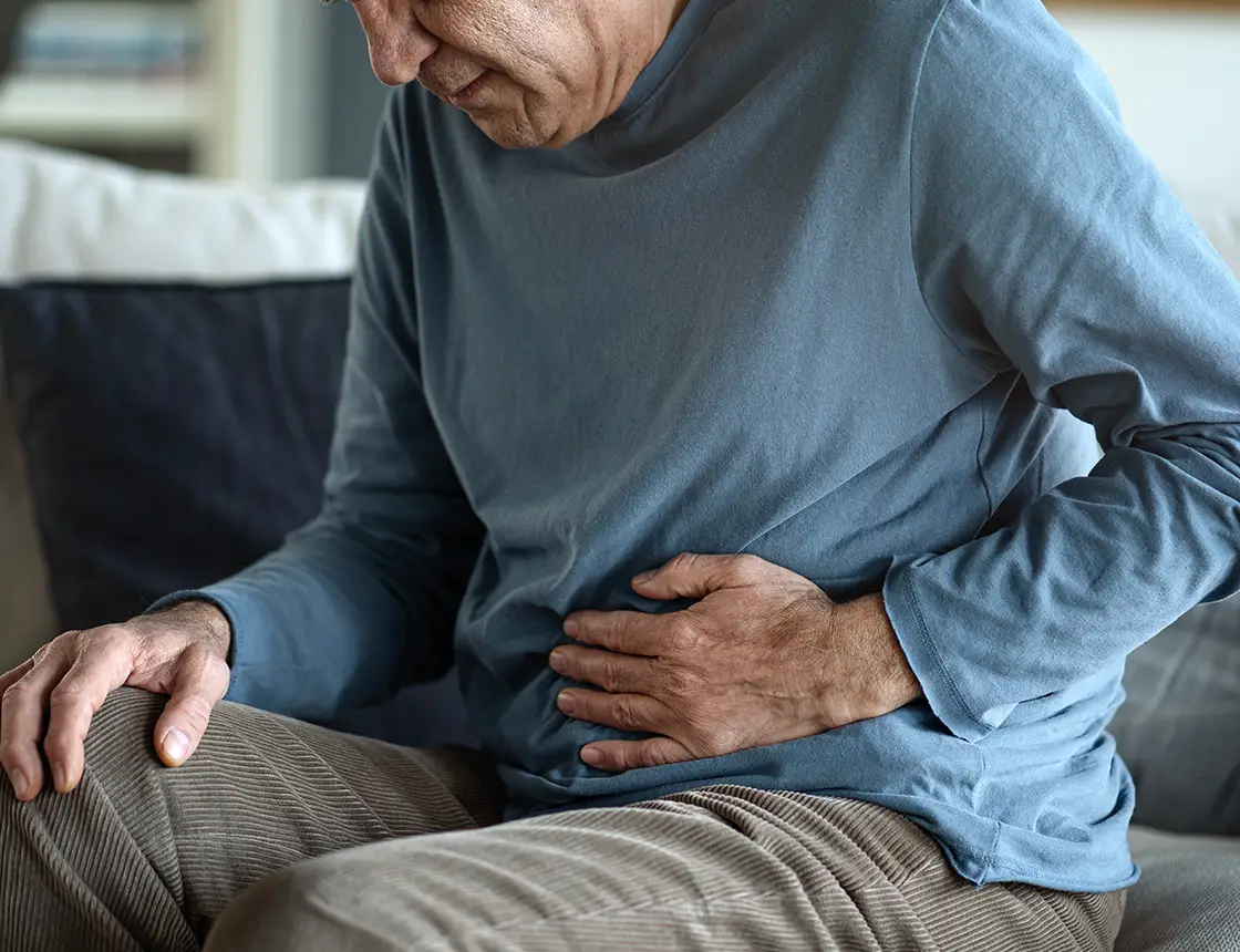 Man sitting on a couch holding his stomach, appearing to have abdominal pain or discomfort.