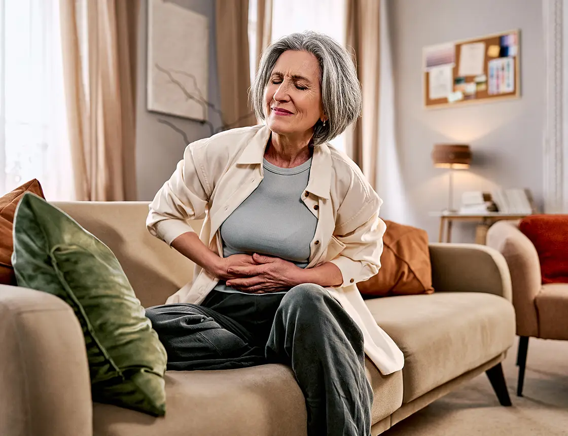 Older woman sitting on sofa clutching stomach with eyes closed in apparent abdominal pain.