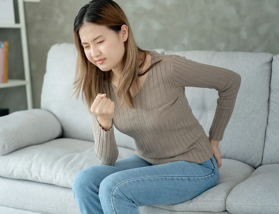 Woman sitting on couch clutching her lower back in pain with eyes closed.