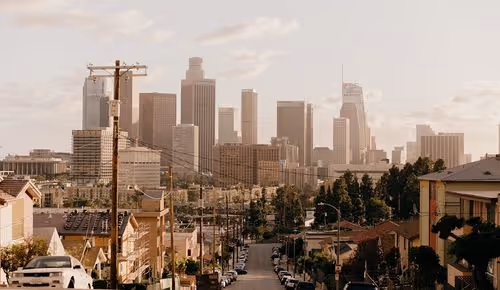 View down a street lined with houses and cars toward a city skyline with tall buildings under a cloudy sky.