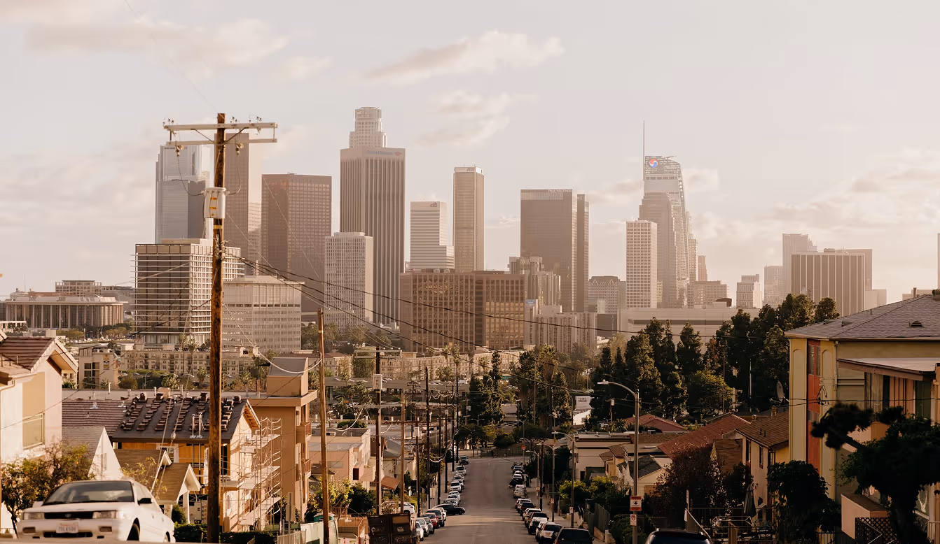 View down a street lined with houses and cars toward a city skyline with tall buildings under a cloudy sky.