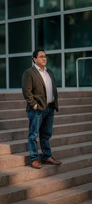 Man wearing a brown jacket, white shirt, and jeans standing on outdoor steps near a large windowed building.