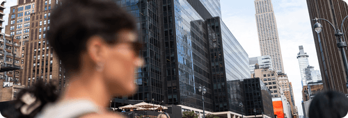 Blurred person with sunglasses in foreground against tall city buildings in a busy urban street.