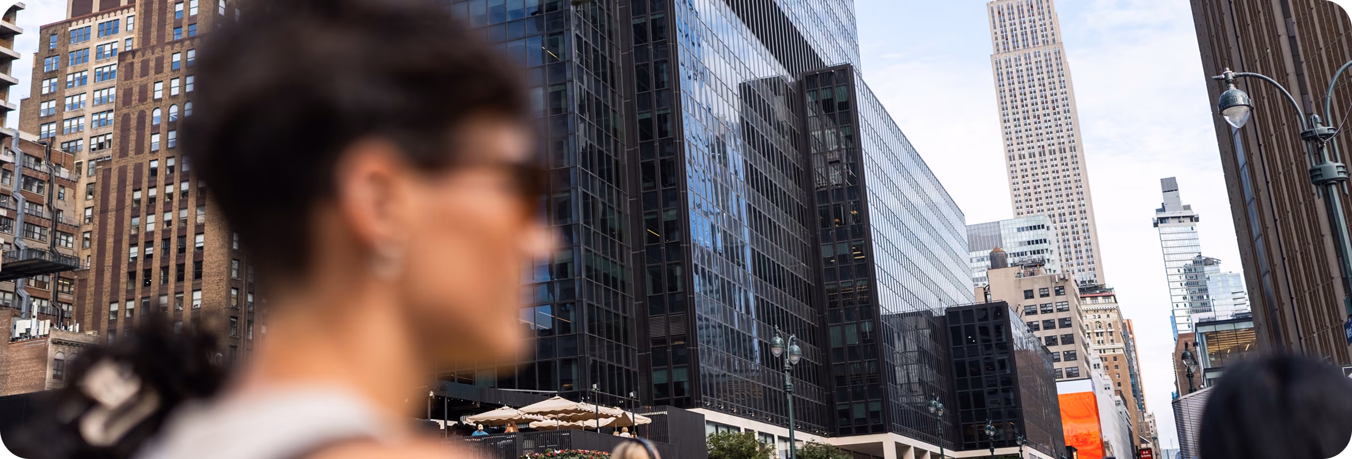 Blurred person with sunglasses in foreground against tall city buildings in a busy urban street.