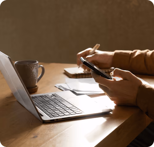 Person at desk using smartphone in one hand and writing notes with a pen on notebook by laptop.