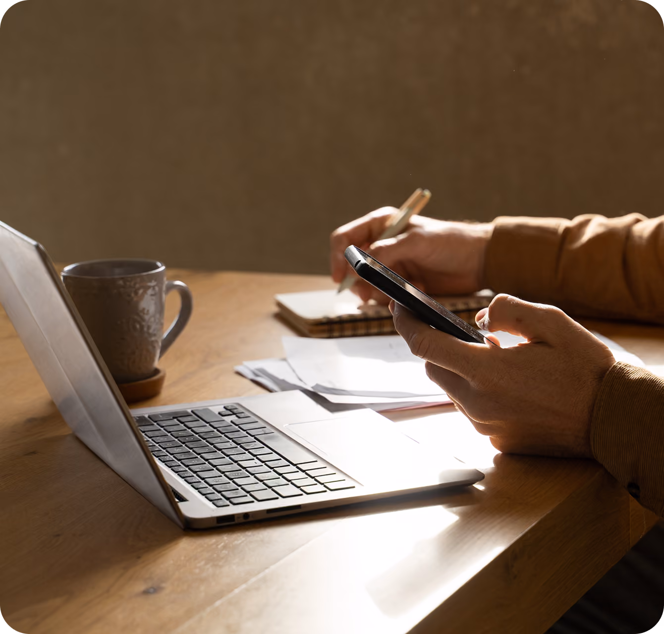 Person at desk using smartphone in one hand and writing notes with a pen on notebook by laptop.