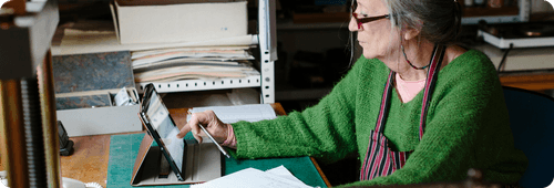Older woman in green sweater using a tablet with a stylus at a desk with papers.
