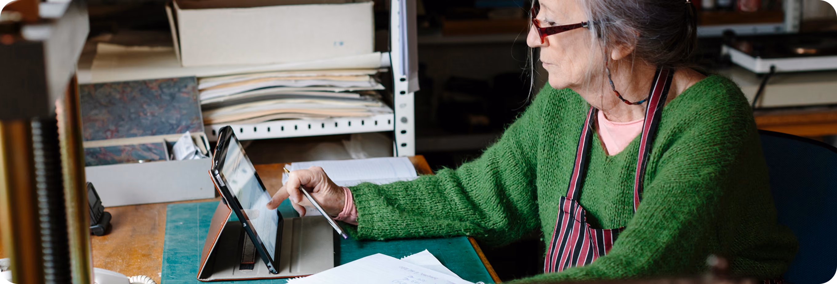 Older woman in green sweater using a tablet with a stylus at a desk with papers.